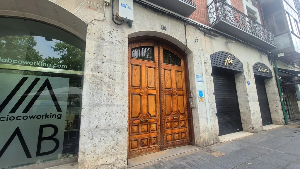 Street view of a building with a large, ornate wooden door, flanked by storefronts with closed metal shutters.