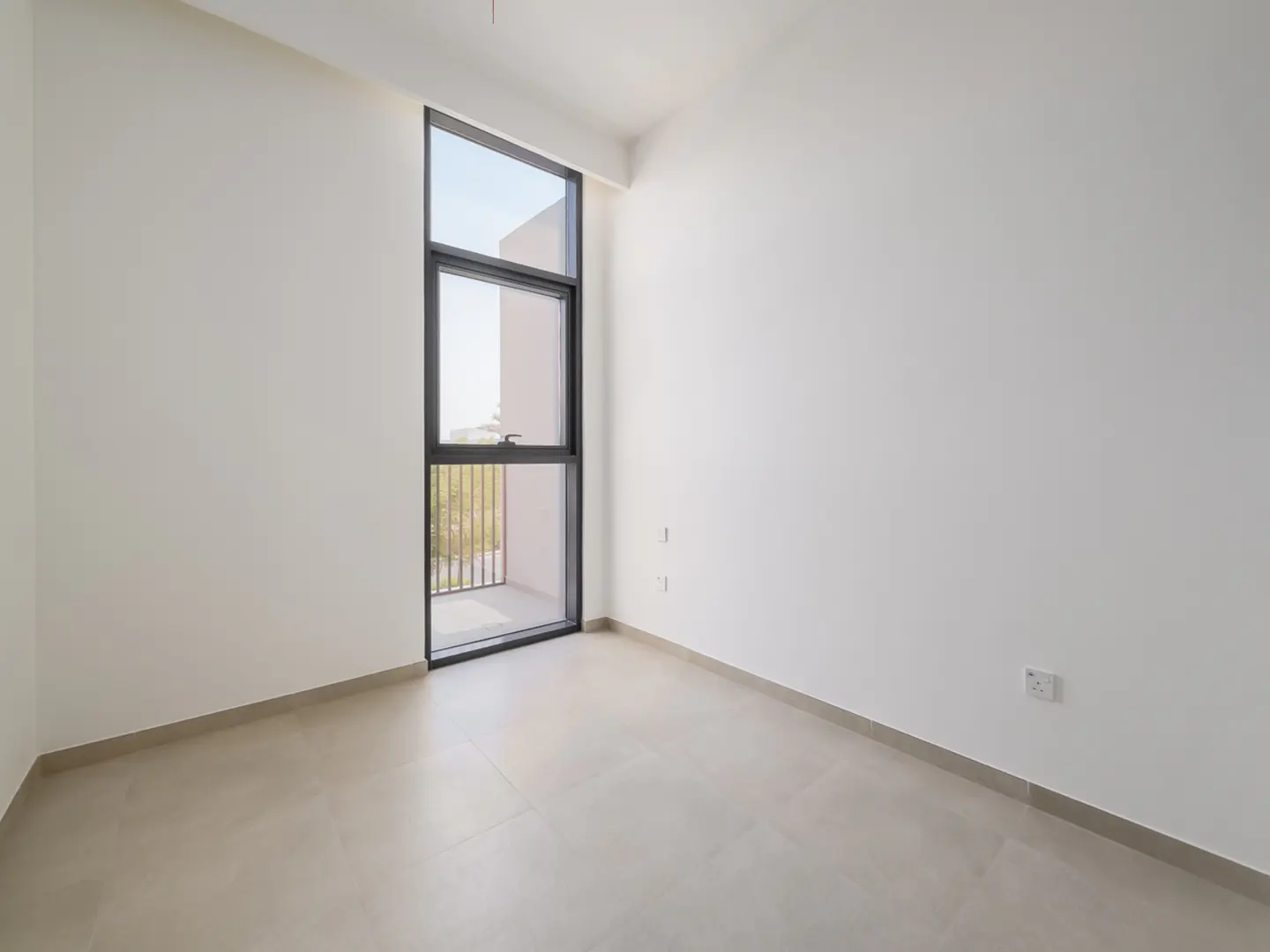 Bright, empty room with white walls and beige tile floor. A tall, black-framed window shows a balcony and greenery outside.