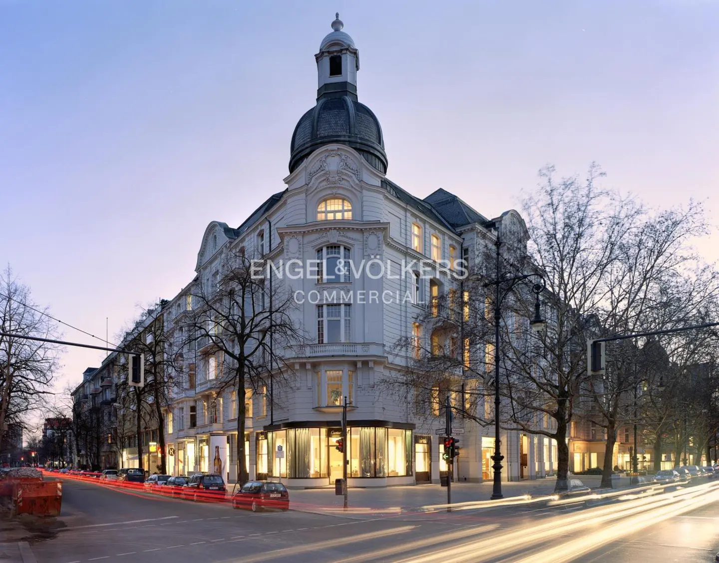 A white, ornate building with a dome at dusk, branded with "Engel & Völkers Commercial," sits at a city intersection with blurred car light trails.