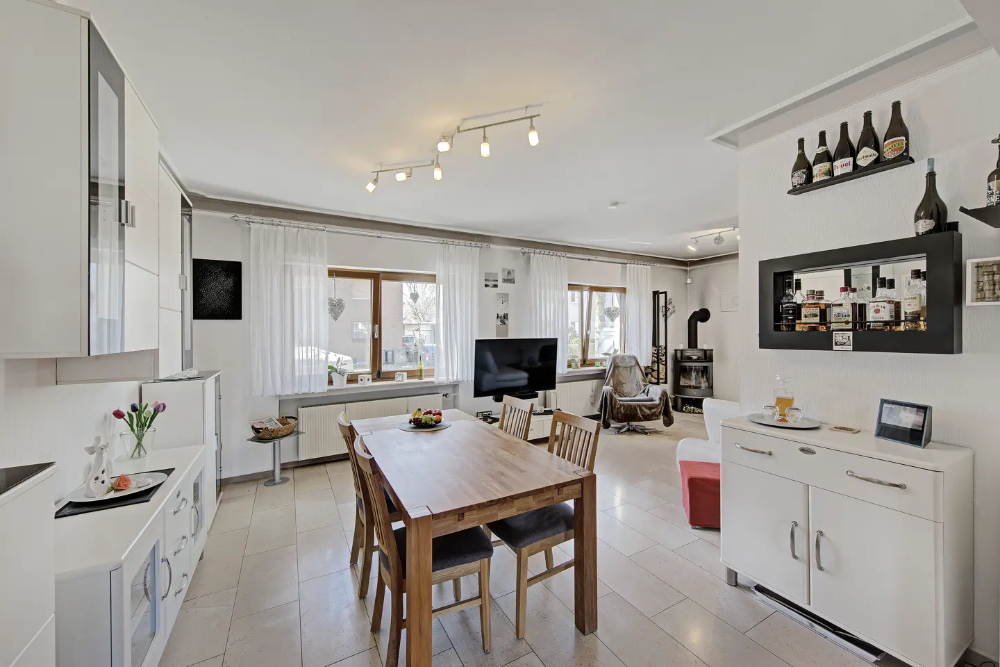 Bright, open-concept living space with a wooden dining table, white cabinets, and a bar area with liquor bottles on shelves.