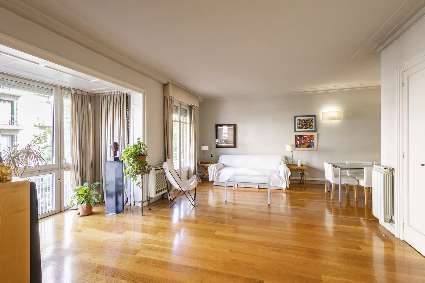 Bright living room with hardwood floors, white sofa, glass table, and large windows with curtains. Plants and artwork add color.