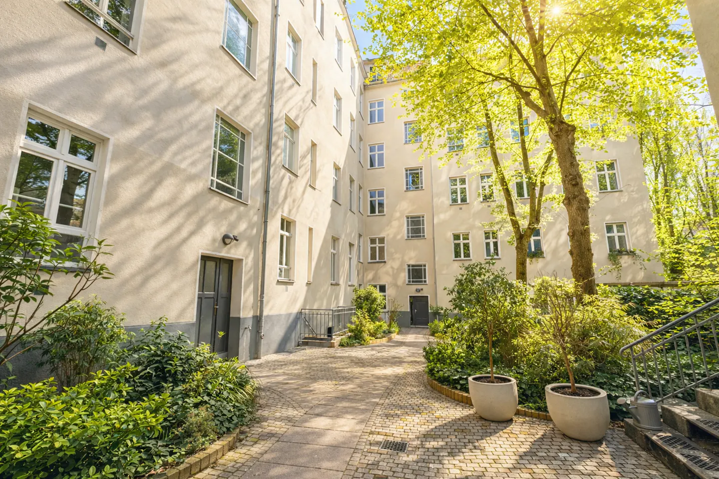 Courtyard view of a multi-story apartment building with green trees and plants. The building is light beige with white framed windows.