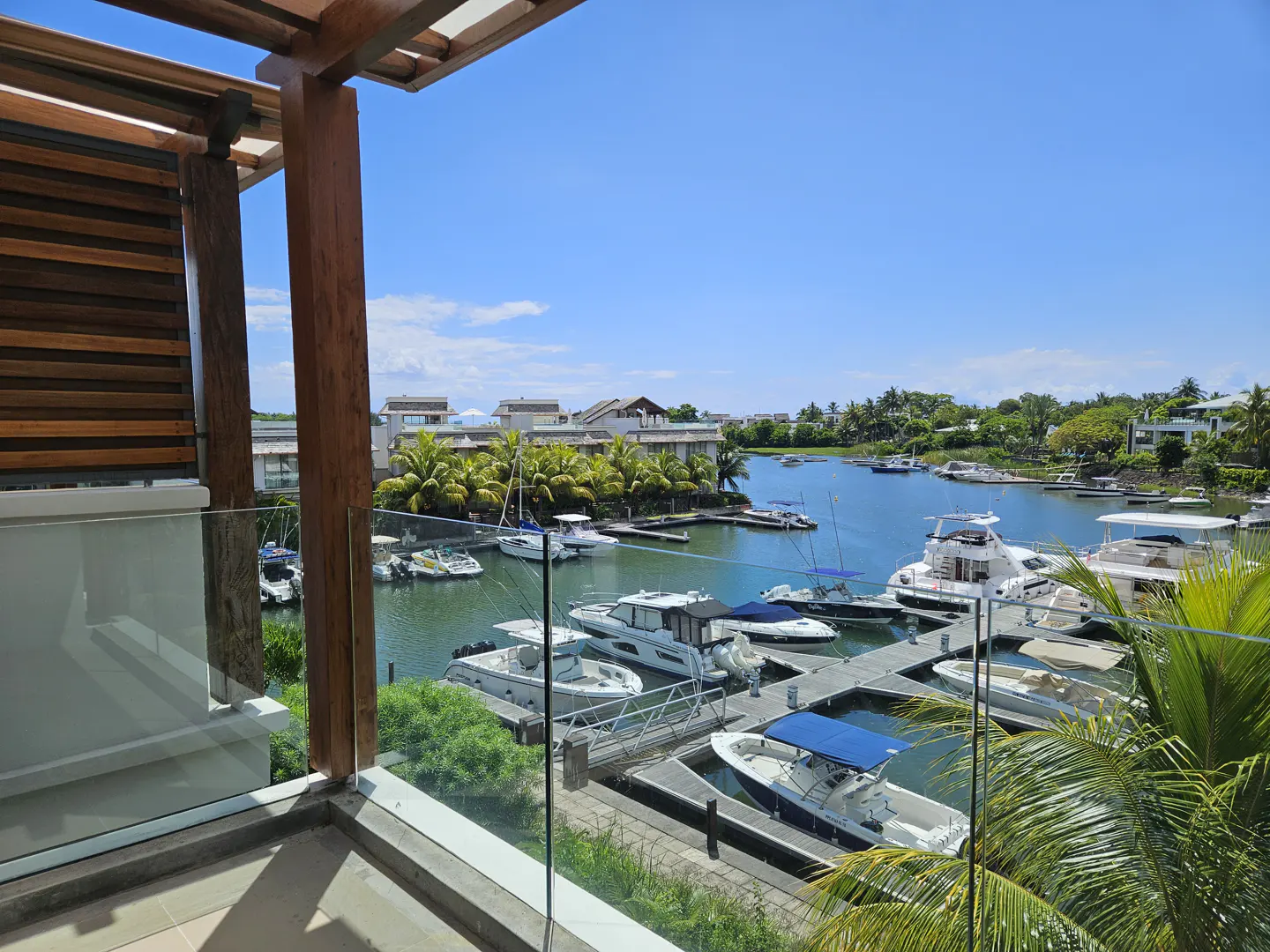 Balcony view of a marina with boats docked on a sunny day. Palm trees and buildings are visible in the background.