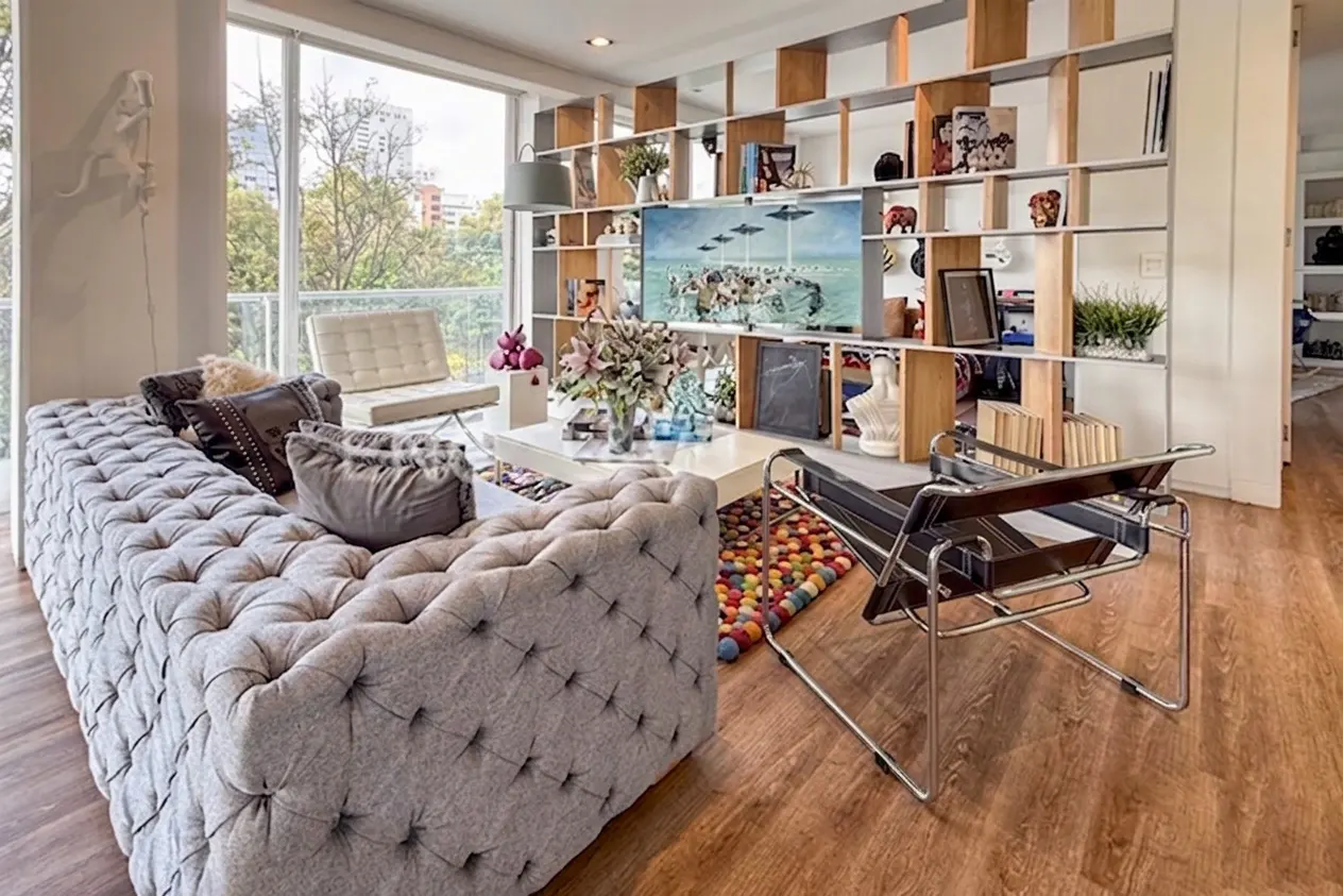 Living room with a gray tufted sofa, white coffee table, and a bookshelf displaying art and decor. A black leather chair sits on a colorful rug.