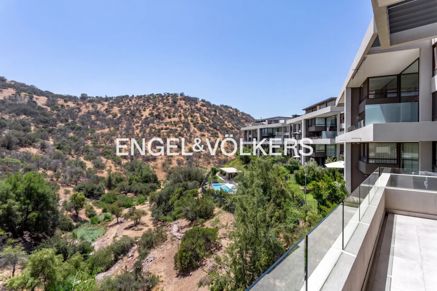 View from a modern balcony overlooking a hillside with trees, a pool, and contemporary buildings under a clear blue sky.