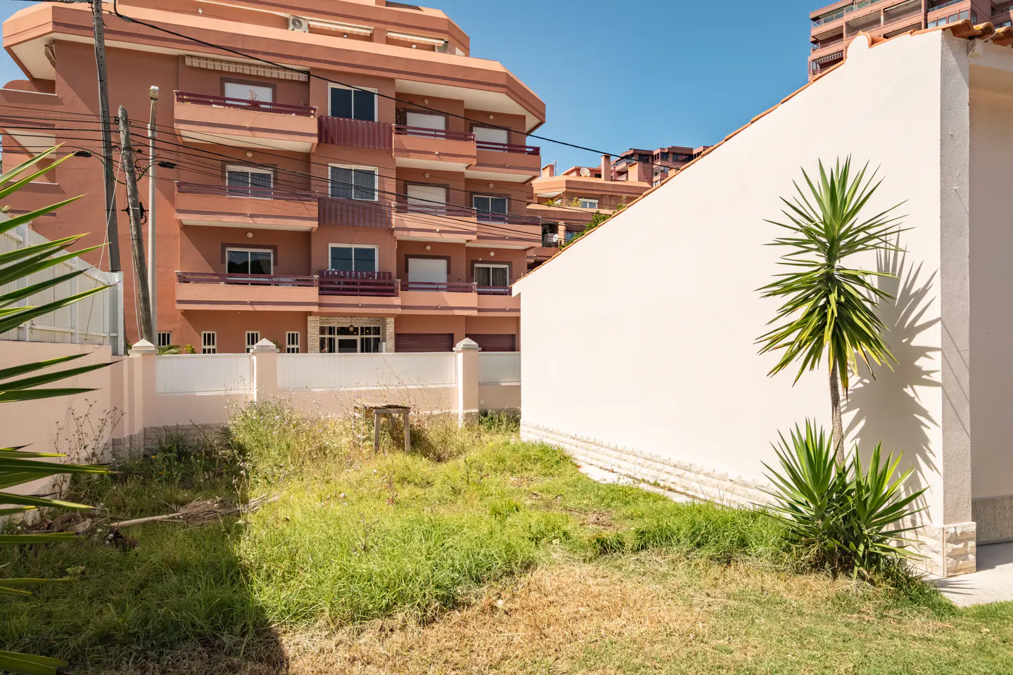 A vacant lot with overgrown grass, a white wall, and a multi-story peach-colored building in the background.
