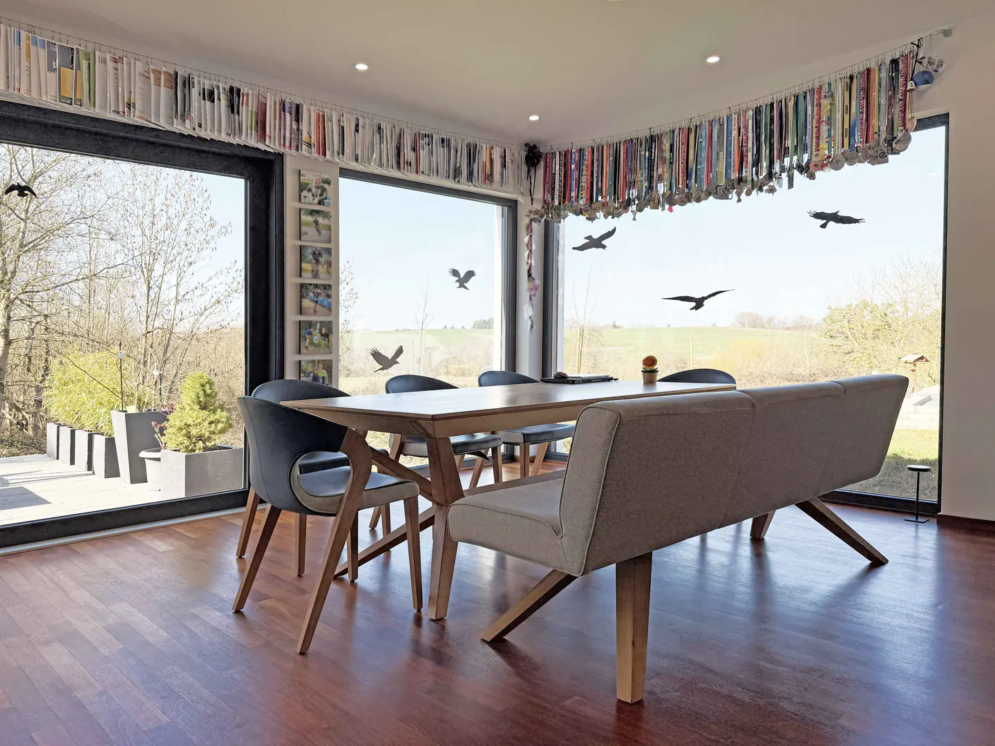 Bright dining room with wood floors, table, chairs, and bench. Large windows overlook a green landscape. Medals and books line the top of the walls.