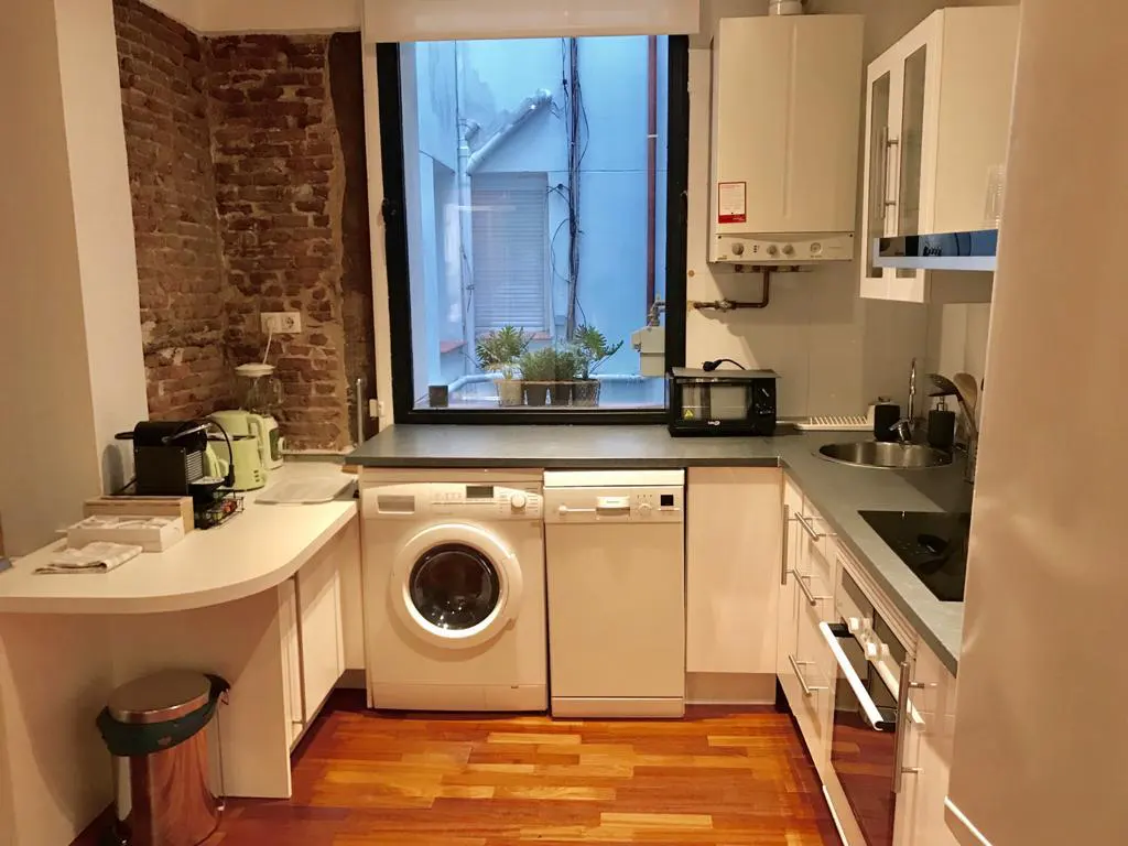 Modern kitchen with white cabinets, wood floors, exposed brick wall, and a window overlooking a small outdoor area.