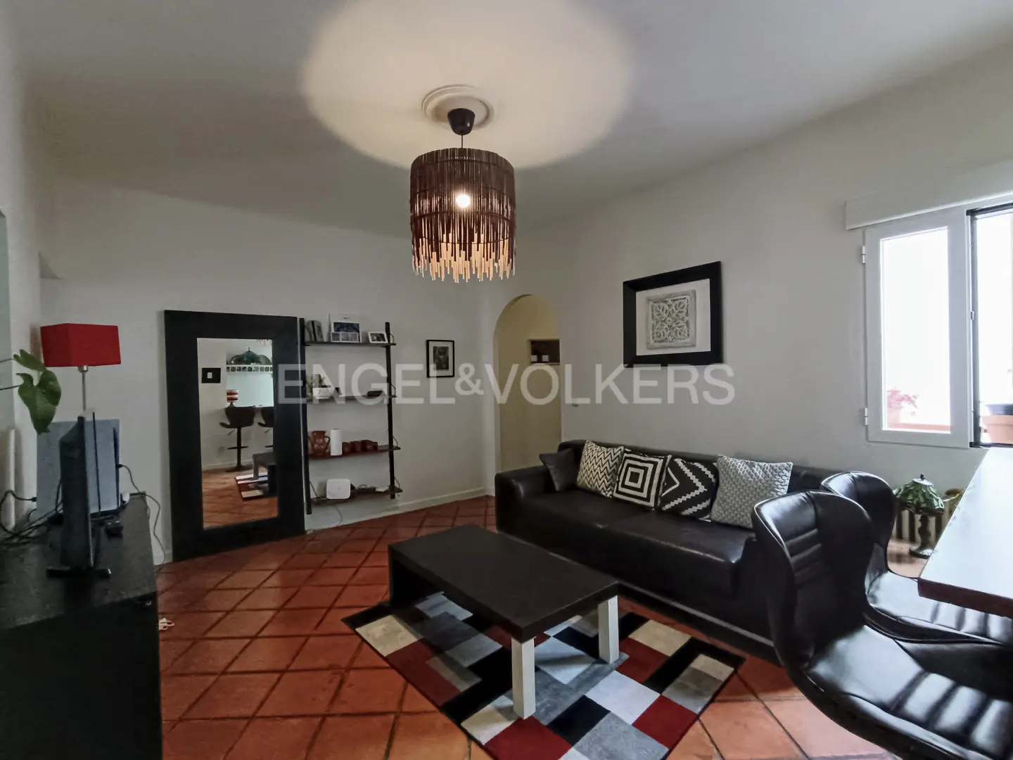 Living room with terracotta tile floors, black leather sofa, patterned rug, coffee table, and a unique fringed chandelier.