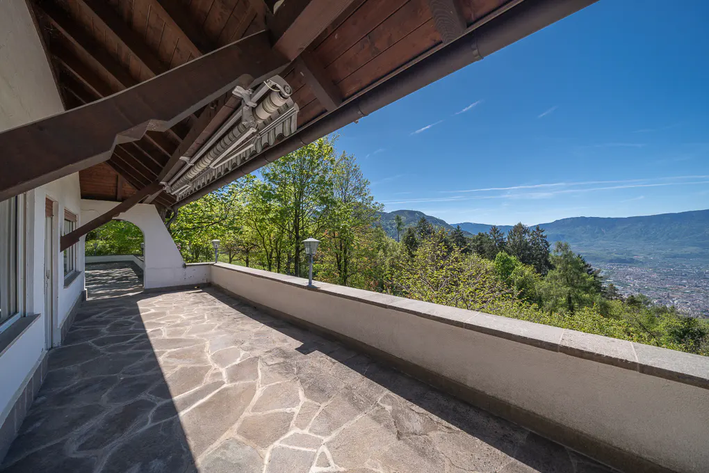 Stone patio with a white wall overlooking a valley with trees and mountains under a blue sky.