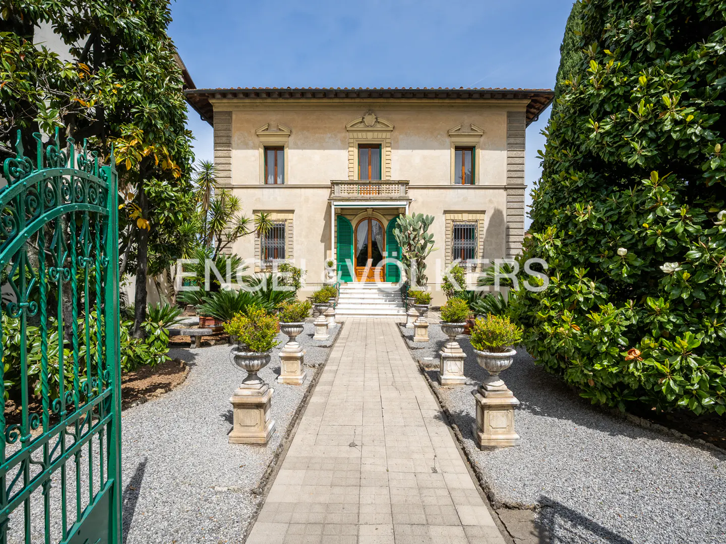 Two-story beige house with green shutters and a stone walkway lined with potted plants. Green gate on the left.