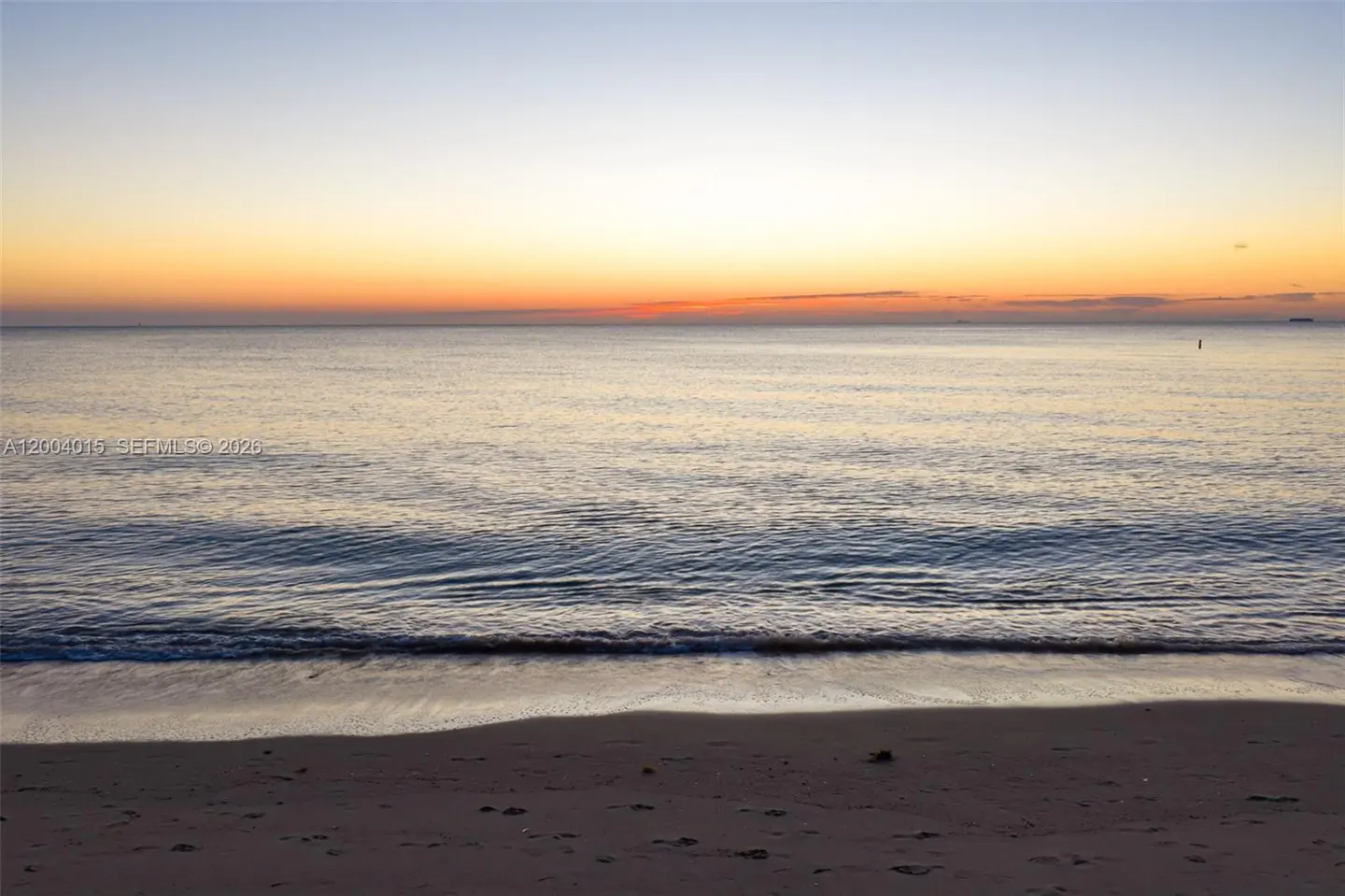 Sunrise over a calm ocean, with a sandy beach in the foreground. The sky is a gradient of orange, yellow, and blue.