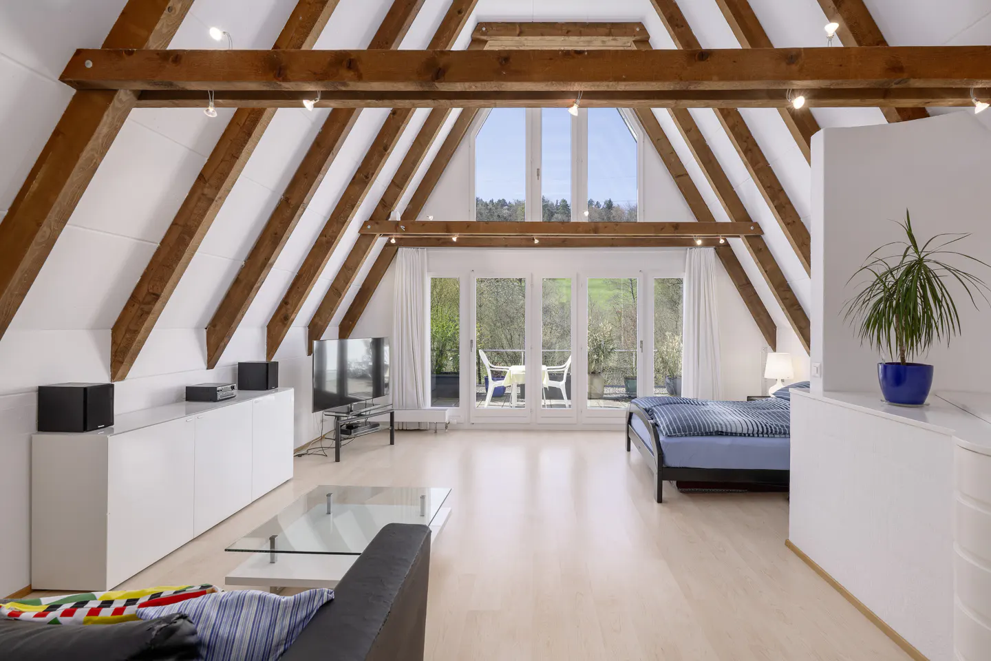 Attic apartment with exposed beams, white walls, and light wood floors. A bed, sofa, and TV furnish the space, with a balcony overlooking greenery.