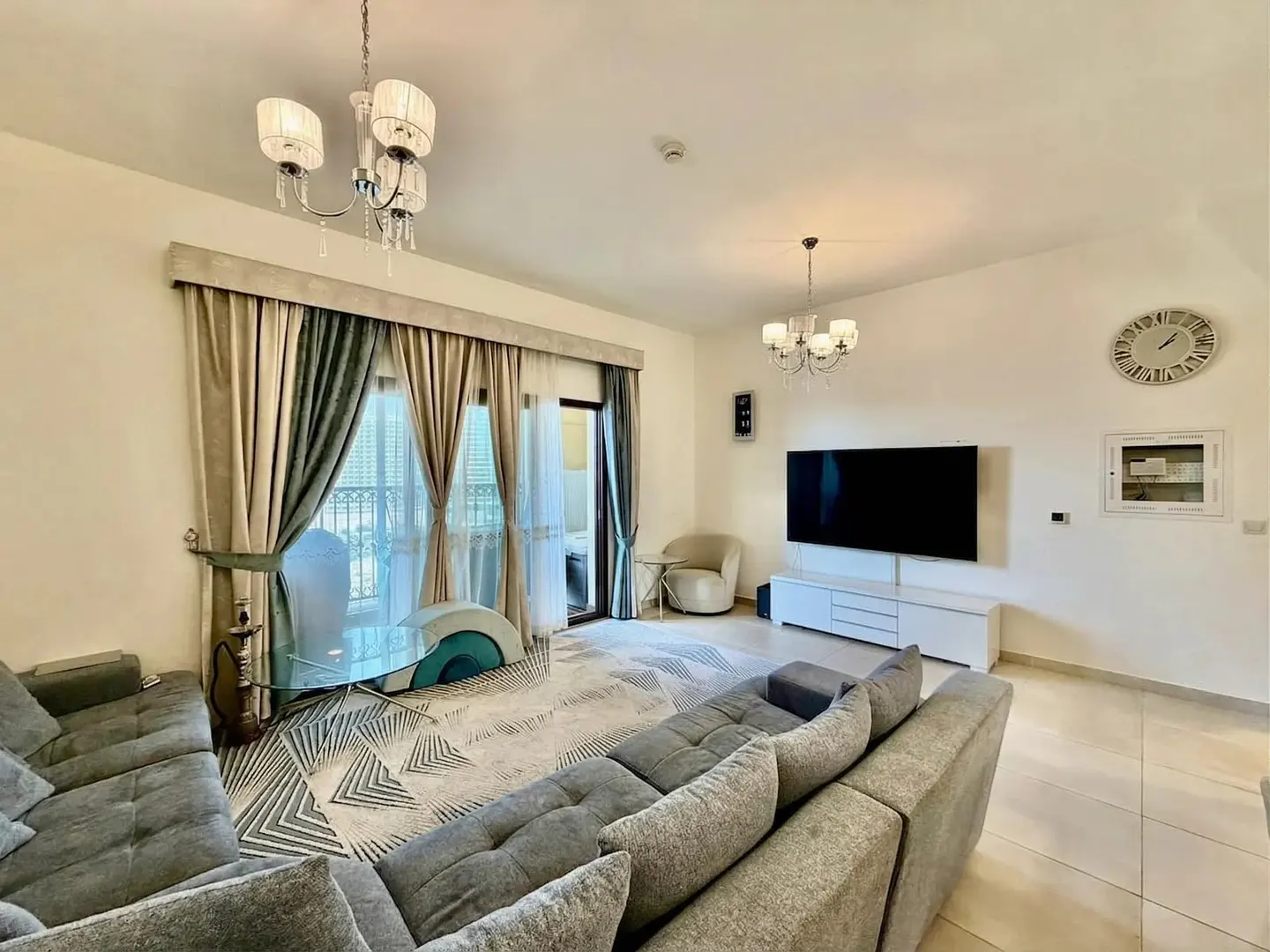 Living room with gray sectional sofa, patterned rug, glass table, TV, and balcony access. Neutral color scheme with chandeliers.