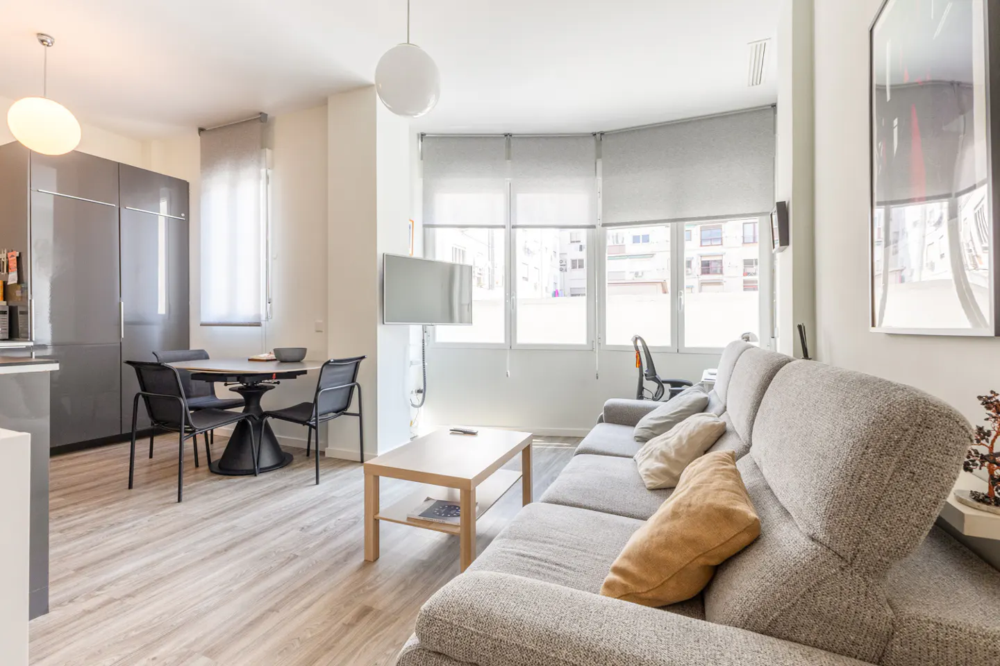 Bright, modern apartment interior with gray sofa, wood floors, dining table, and large windows with roller shades.