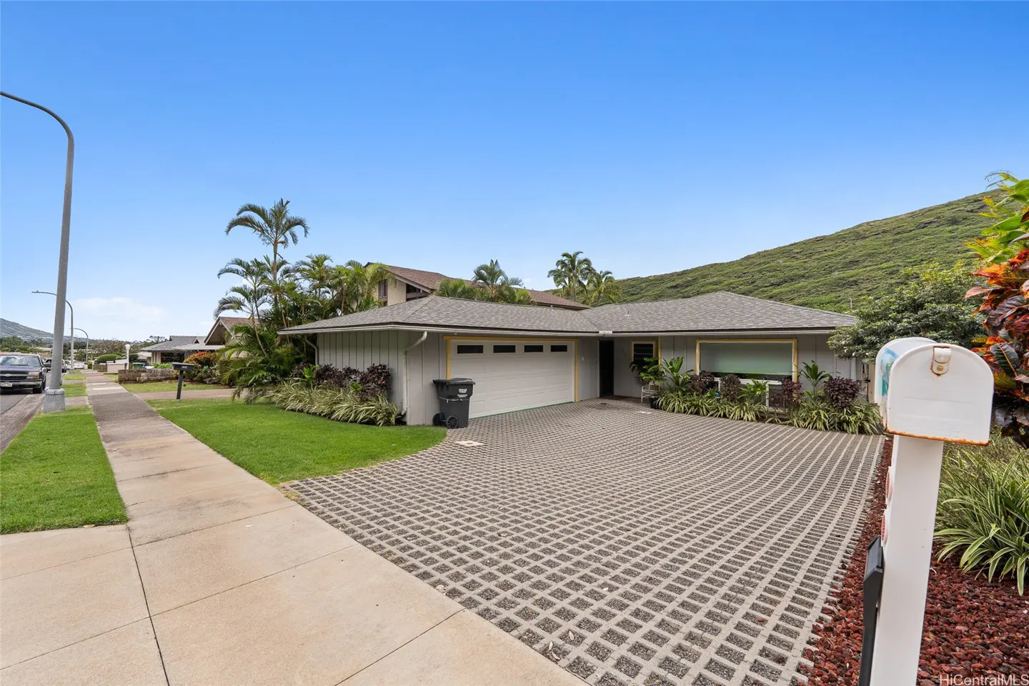 A single-story gray house with a white garage door and a patterned driveway on a sunny day.