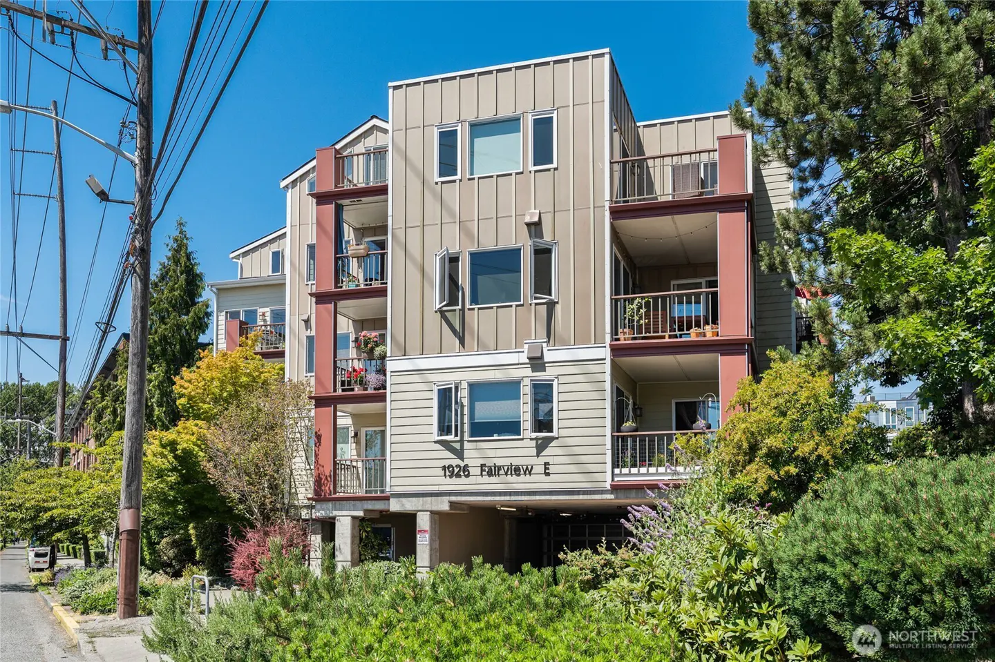 Modern three-story apartment building at 1926 Fairview E with tan siding, red accents, and balconies under a clear blue sky.