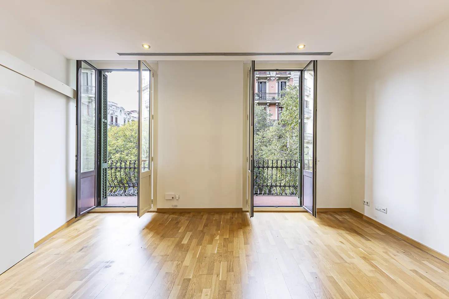 Empty room with light wood floors, white walls, and two open balcony doors overlooking trees and city buildings.