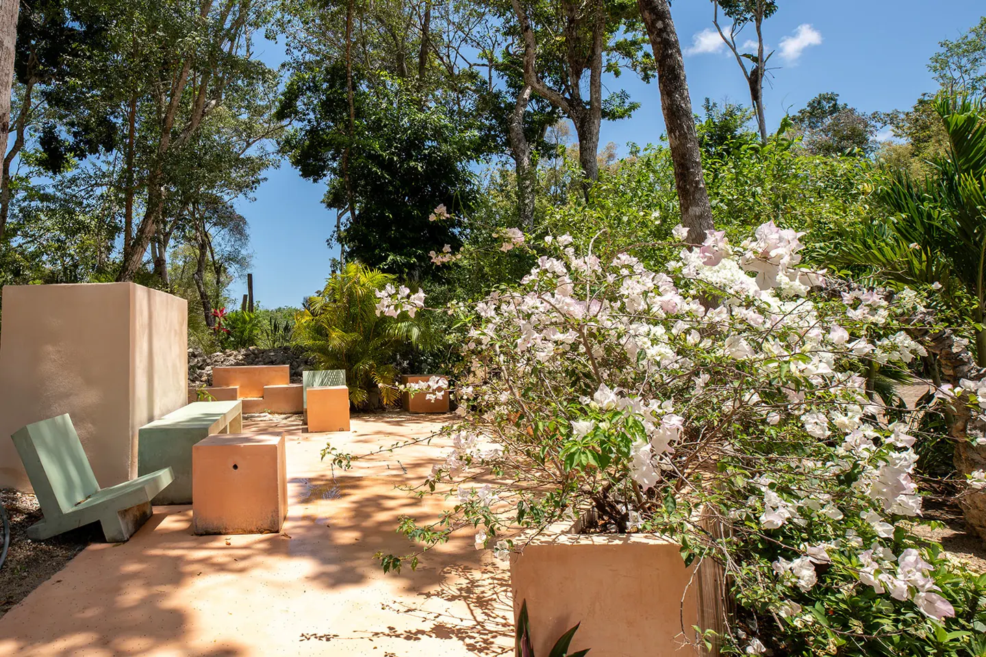 Outdoor patio with concrete furniture, including a green chair, and a large white flowering bush in a pot. Lush green trees in the background.