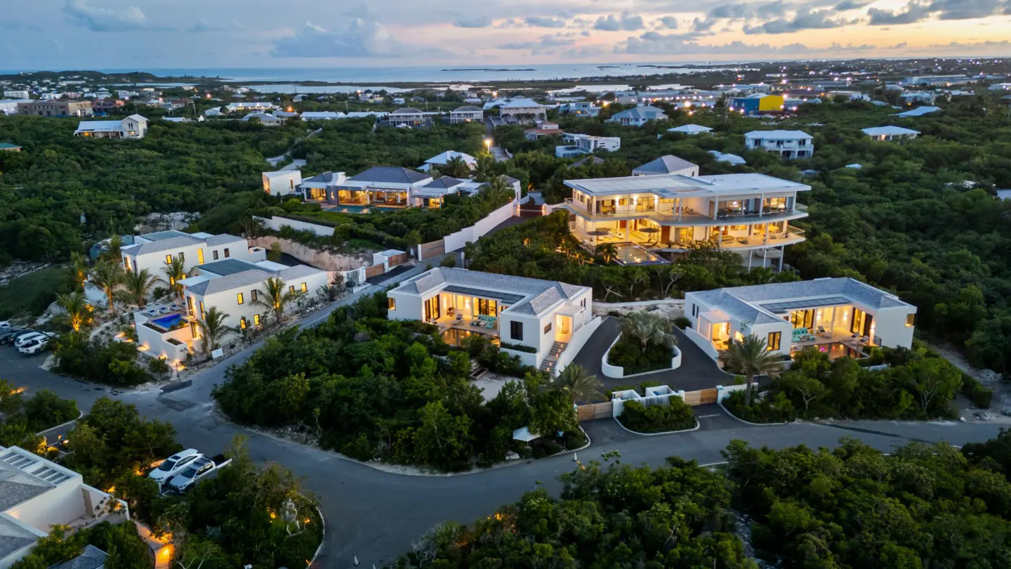 Aerial view of luxury white villas nestled in lush green landscape near the ocean at dusk. Warm lights glow from the homes.