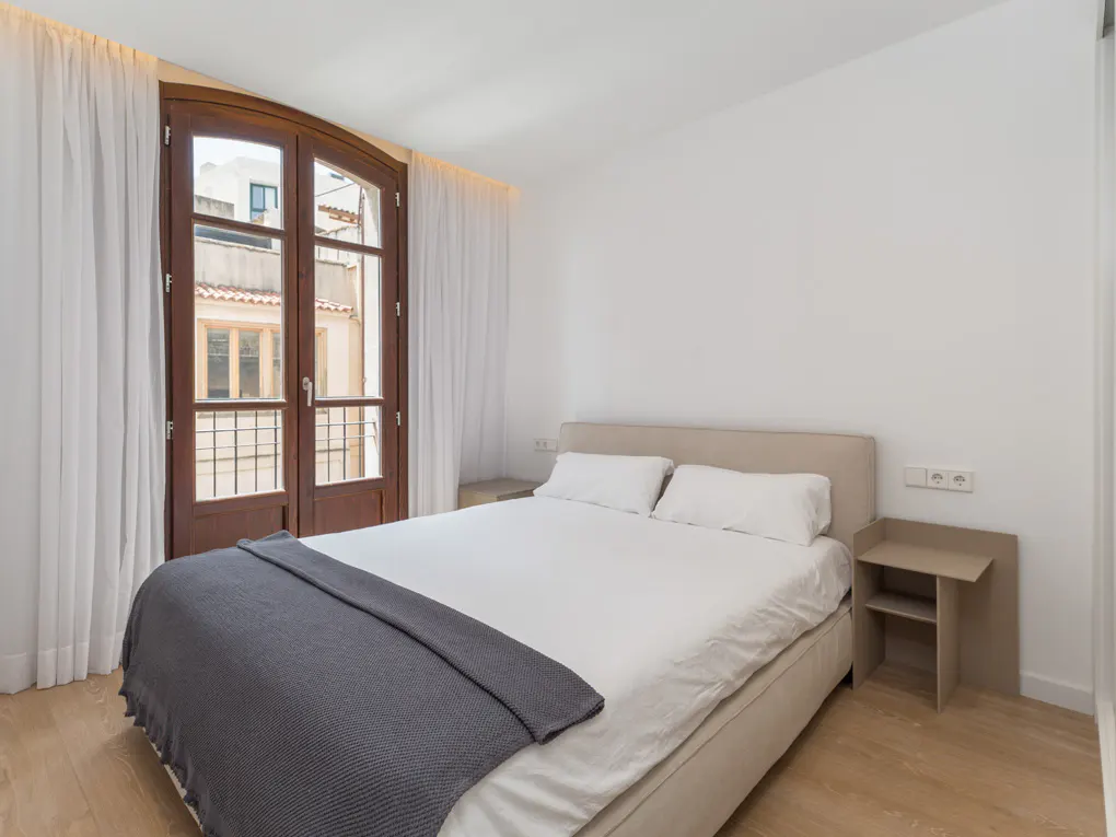 Bedroom with white walls, wood floor, and a bed with white linens and a gray blanket. A wooden door with white curtains leads to a balcony.