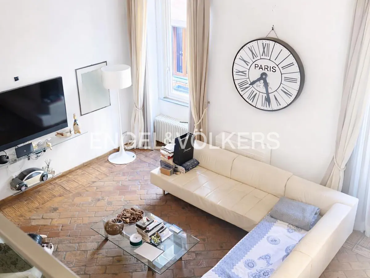 High-angle shot of a living room with white walls, brick floors, and a cream-colored sofa. A large Paris clock hangs on the wall.