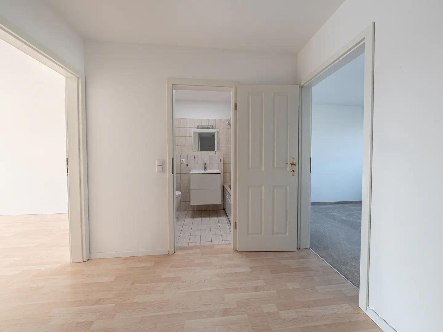 An interior shot of a room with light wood floors and three open doorways. One leads to a bathroom with a white vanity, another to a room with gray carpet.