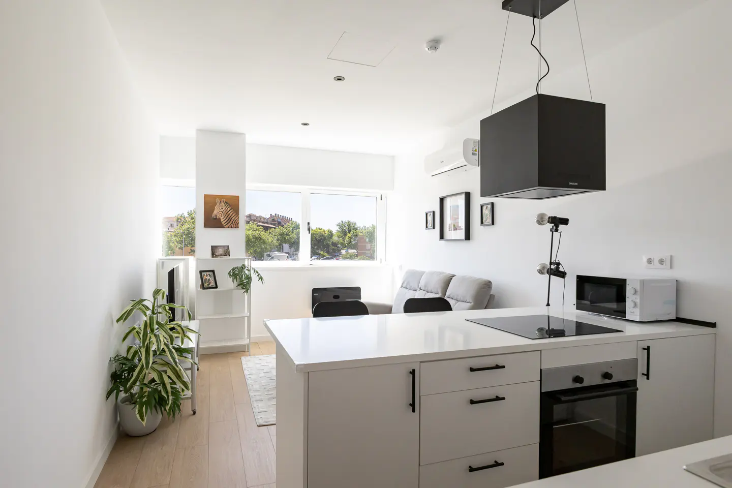 Bright, modern studio apartment with white walls, kitchen island, and living area. Large windows offer a view of trees and buildings.
