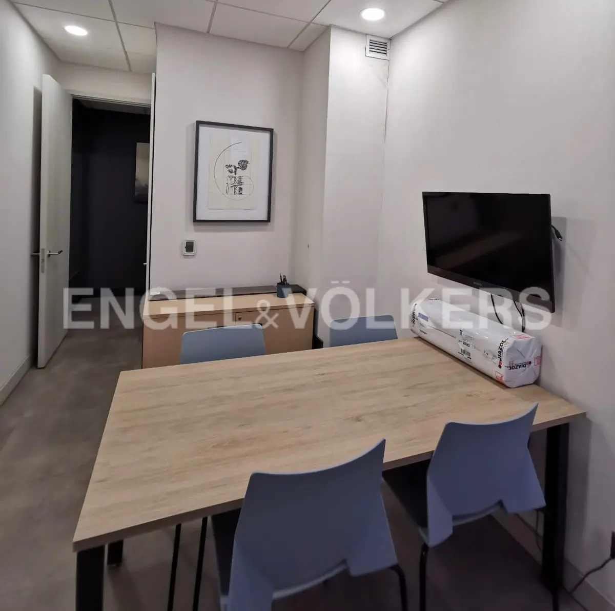 Office space with a light wood table, blue chairs, and a TV mounted on the wall. A framed picture hangs above a wooden cabinet.