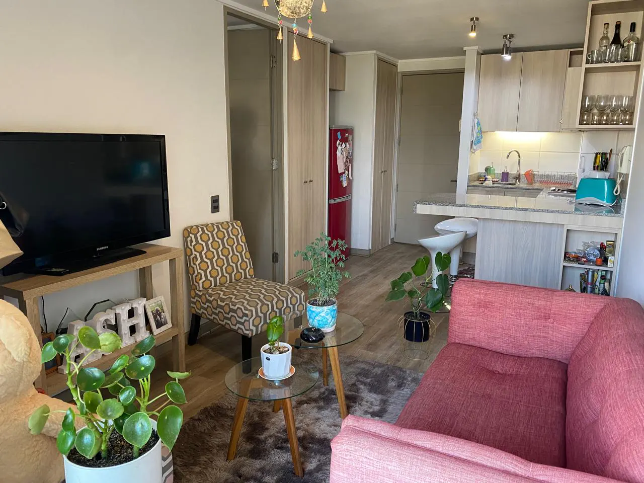 A cozy living room with a TV, patterned chair, pink sofa, and a small kitchen area with a red refrigerator.