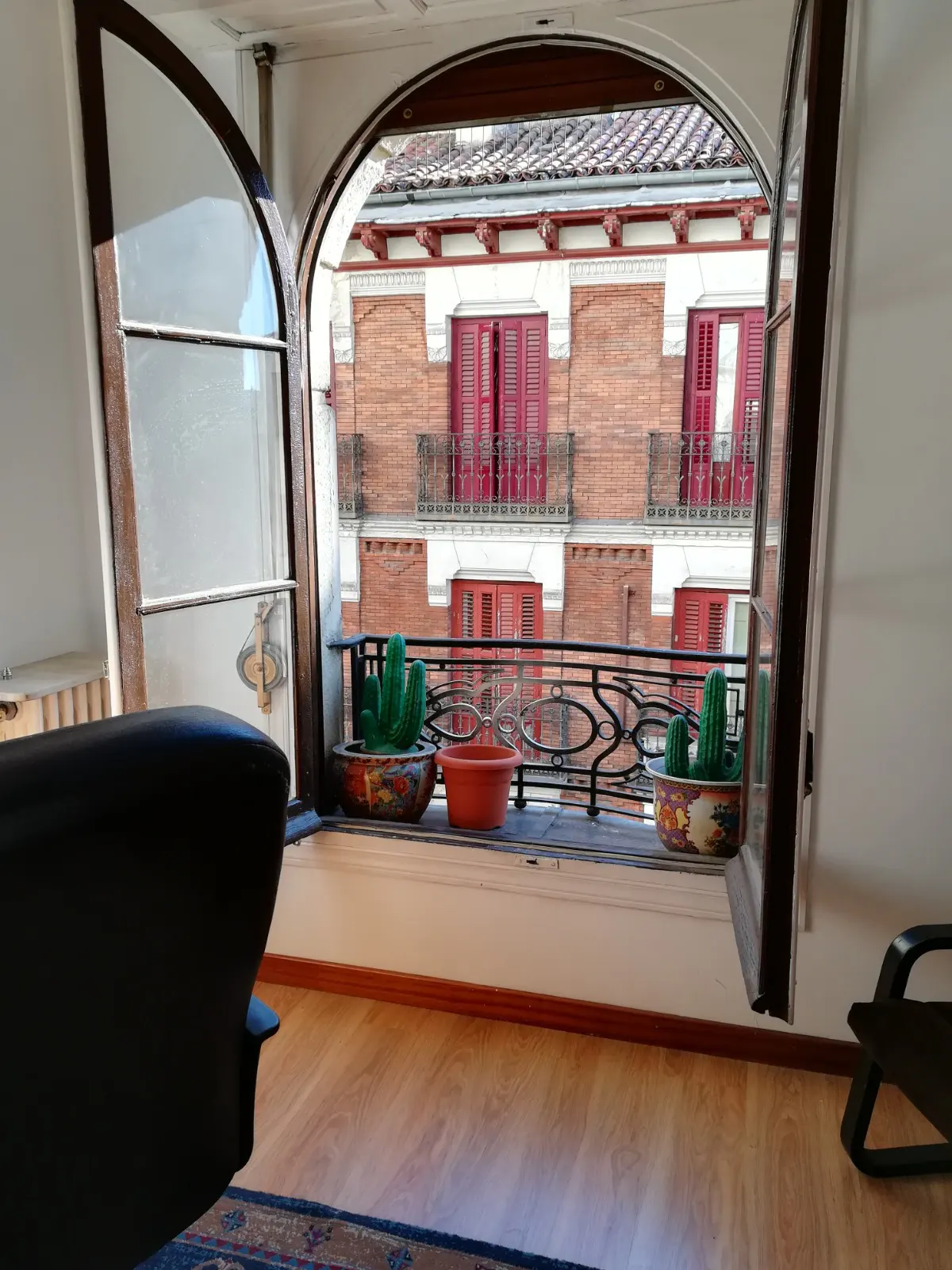 View through arched window with open panes, showing brick building with red shutters and potted cacti on balcony.