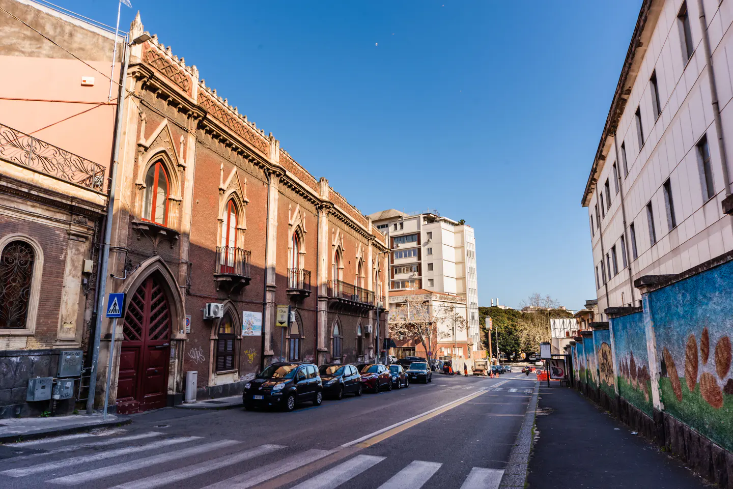 Street view of Catania, Italy, with parked cars, old brick buildings, and a blue sky.