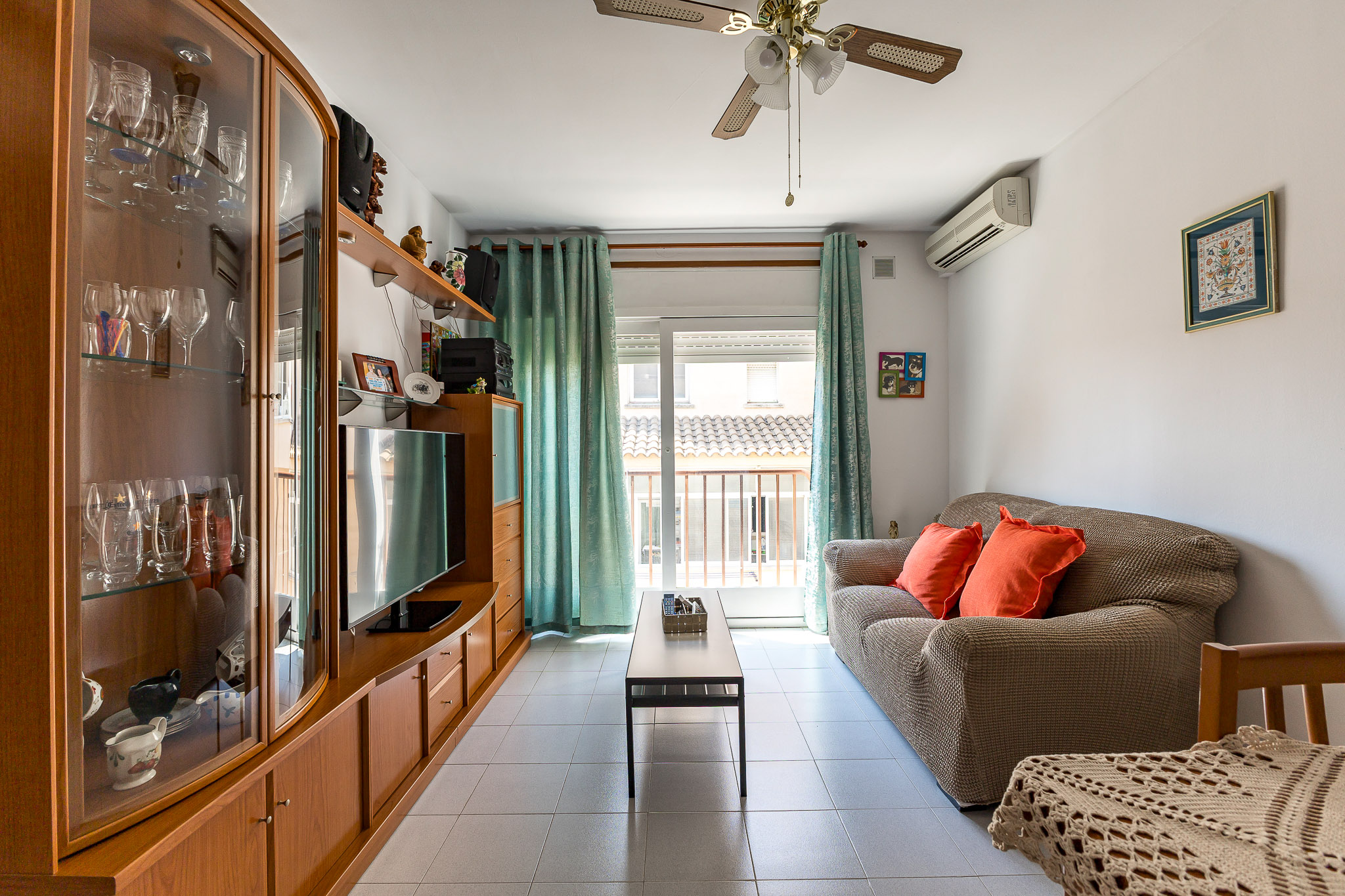 Living room with a beige sofa, orange pillows, wooden media unit, glass display cabinet, and a balcony with teal curtains.