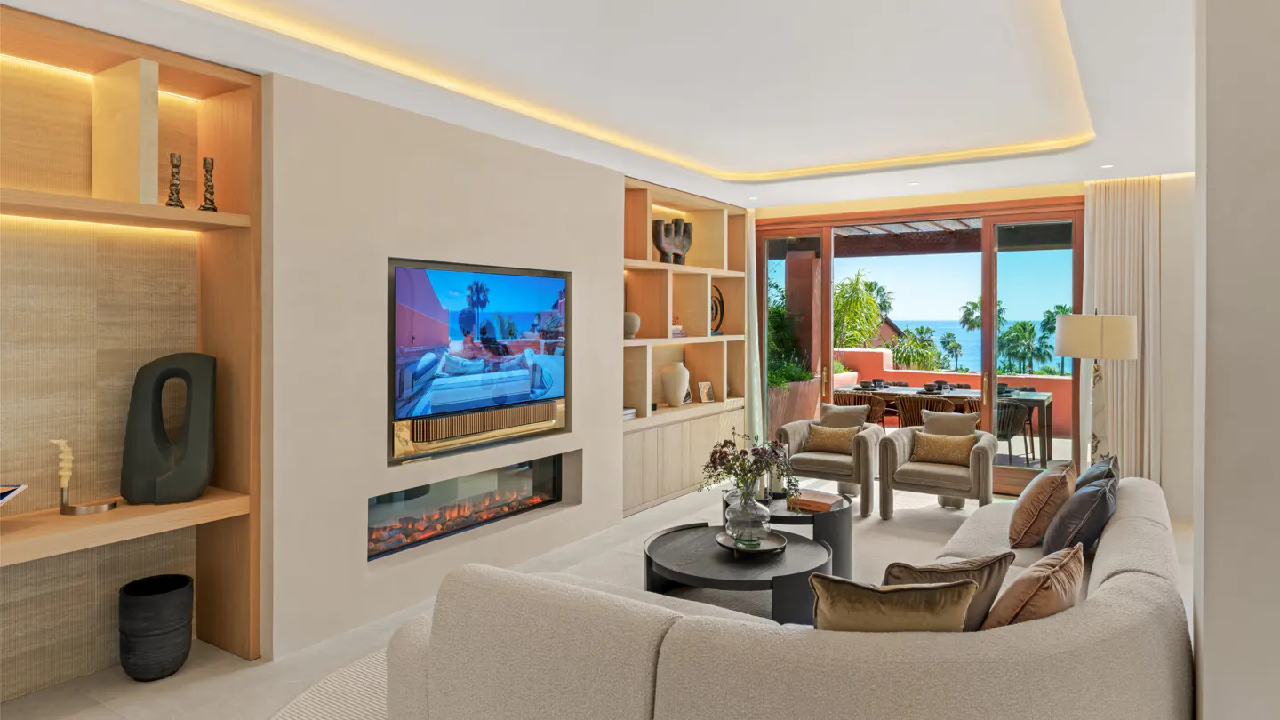 Bright living room with a beige sofa, armchairs, and a TV above a fireplace. A balcony overlooks the ocean with palm trees.