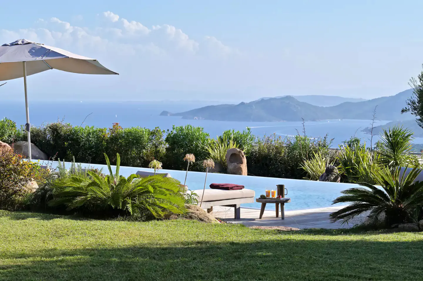 Outdoor infinity pool with lounge chair, drinks, and ocean view. Green lawn and plants surround the pool. Blue sky and mountains in the background.