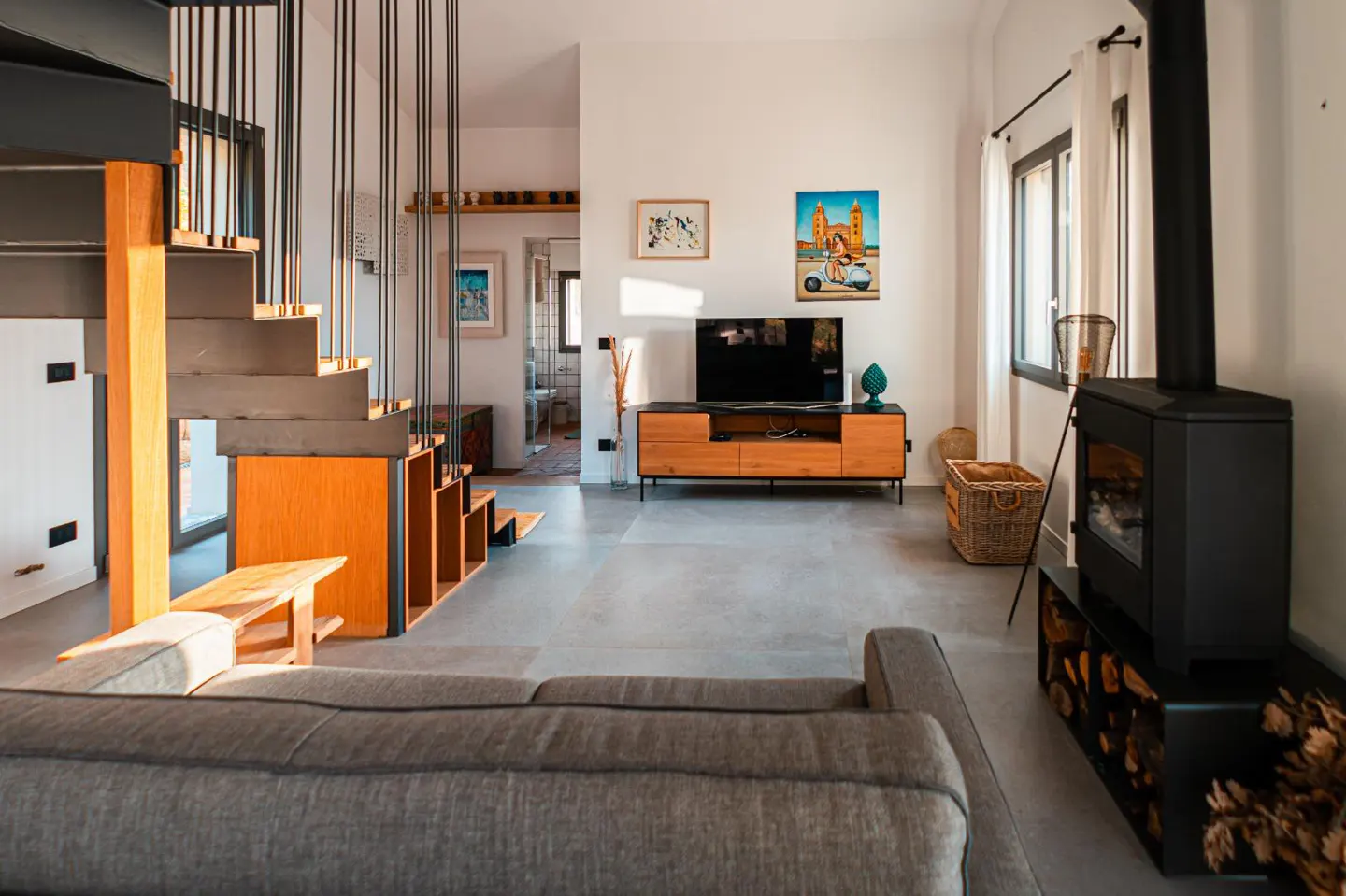 Living room with gray sofa, wood-burning stove, TV, and open staircase. The walls are white, and the floor is gray. There are paintings on the wall.