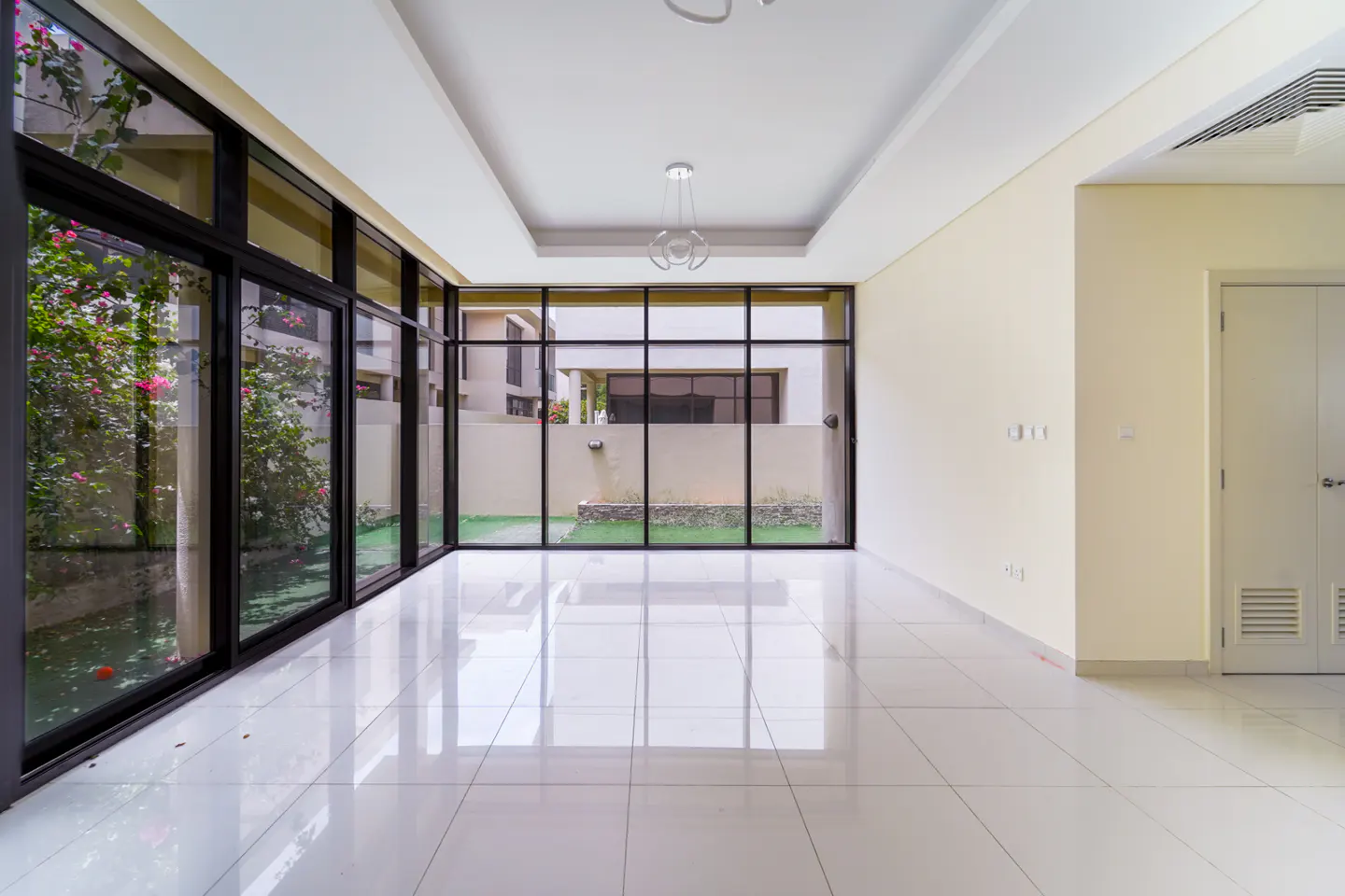 Bright, empty room with white tile floor and large windows. Outside, greenery and a building are visible. A modern chandelier hangs from the ceiling.