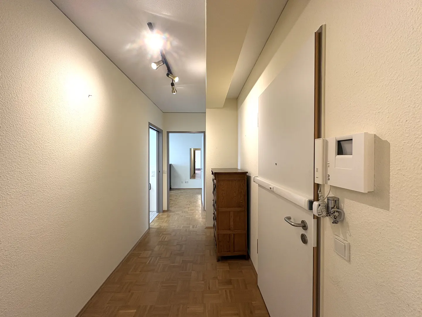 Hallway view with parquet flooring, white walls, and a wooden dresser. A white door with an intercom system is on the right.