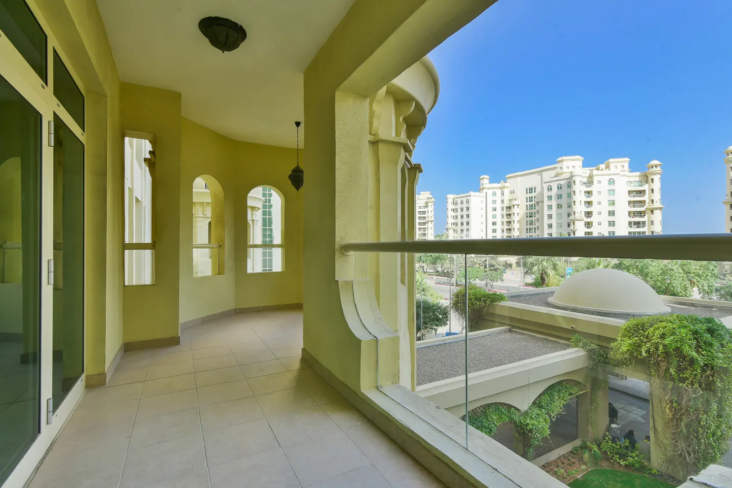 Balcony view of a yellow building with arched windows and a glass railing overlooking a courtyard with a dome and greenery.