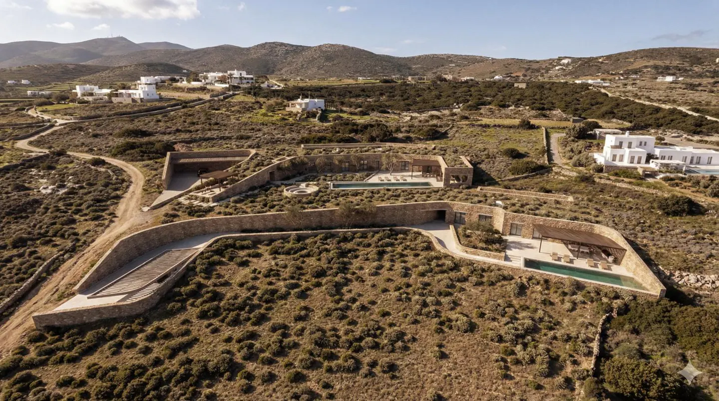 Aerial view of a modern stone house with a pool, nestled in a dry, hilly landscape under a blue sky.