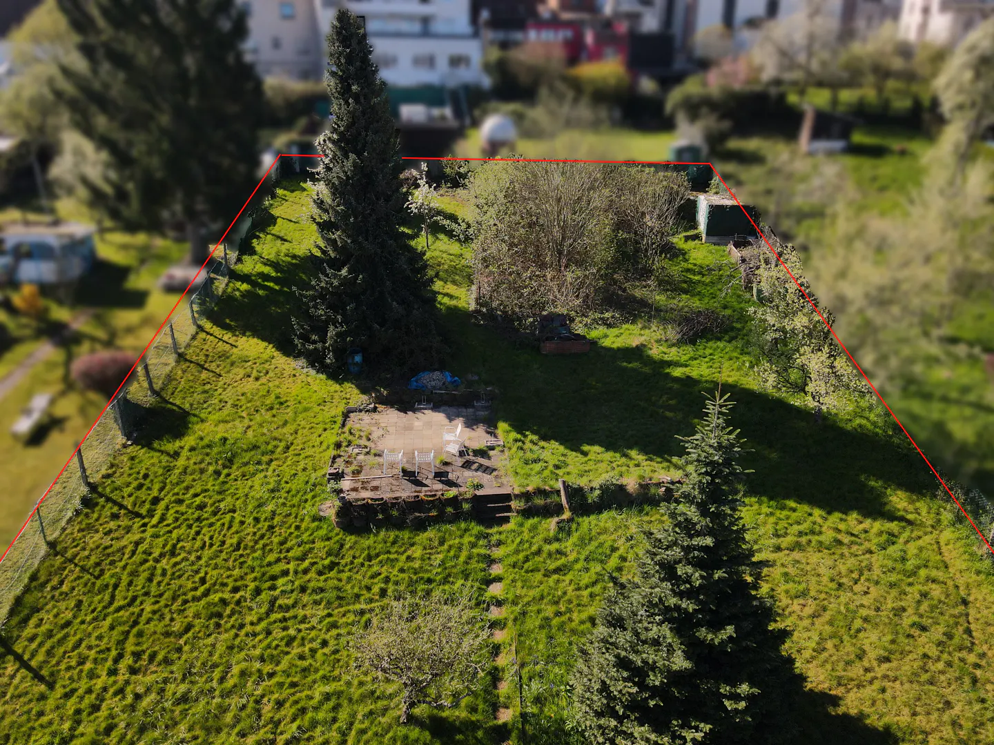 Aerial view of a grassy property outlined in red, featuring trees, a patio with chairs, and a fence.