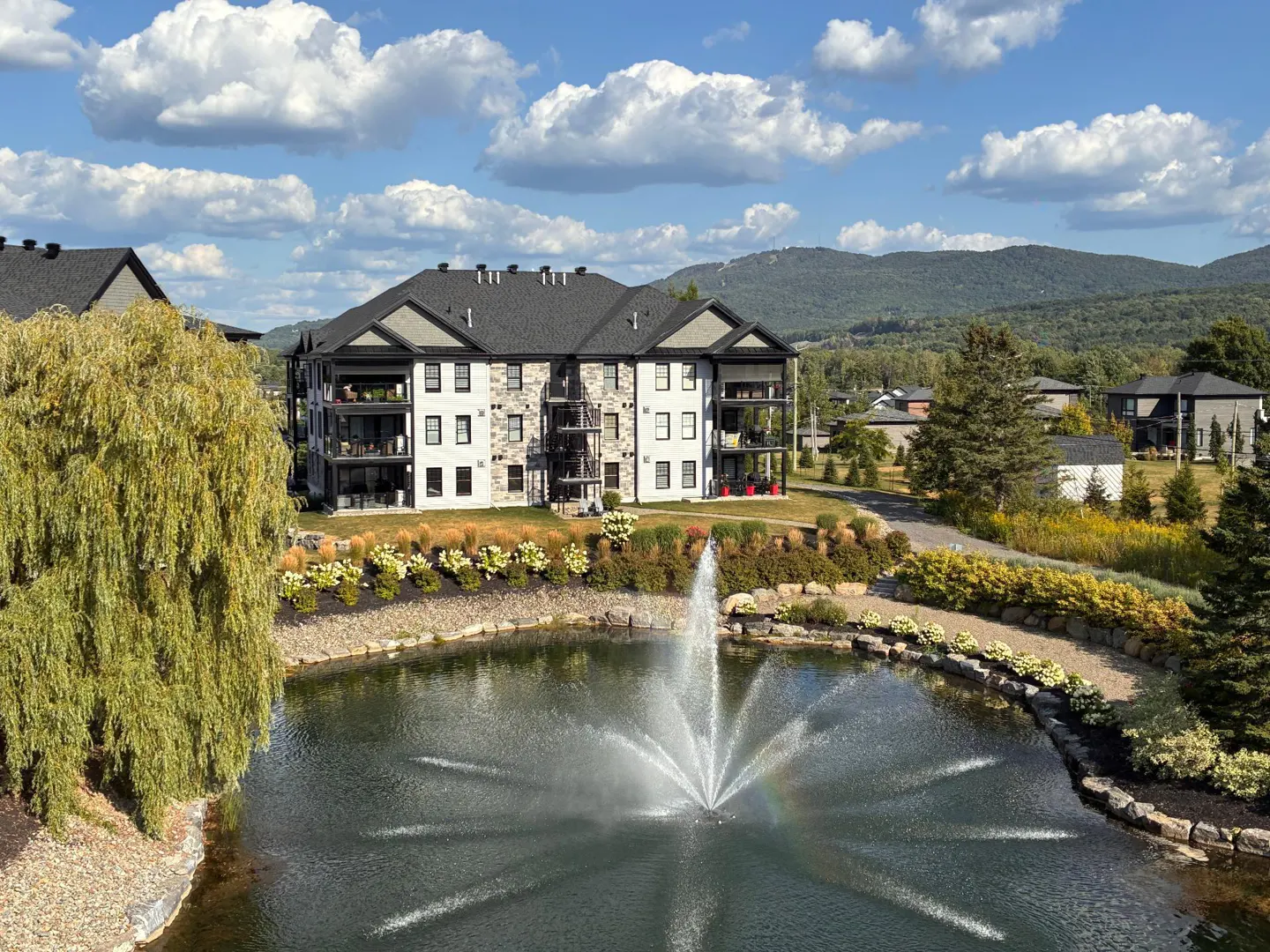 Exterior view of a white and gray apartment building with balconies, a pond with a fountain, and mountains in the background.