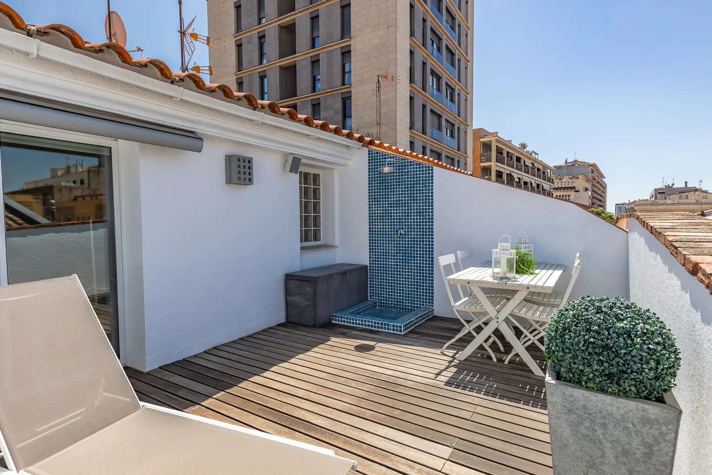 Rooftop terrace with wooden deck, white walls, and city views. Features a blue-tiled shower, white table with chairs, and potted plant.