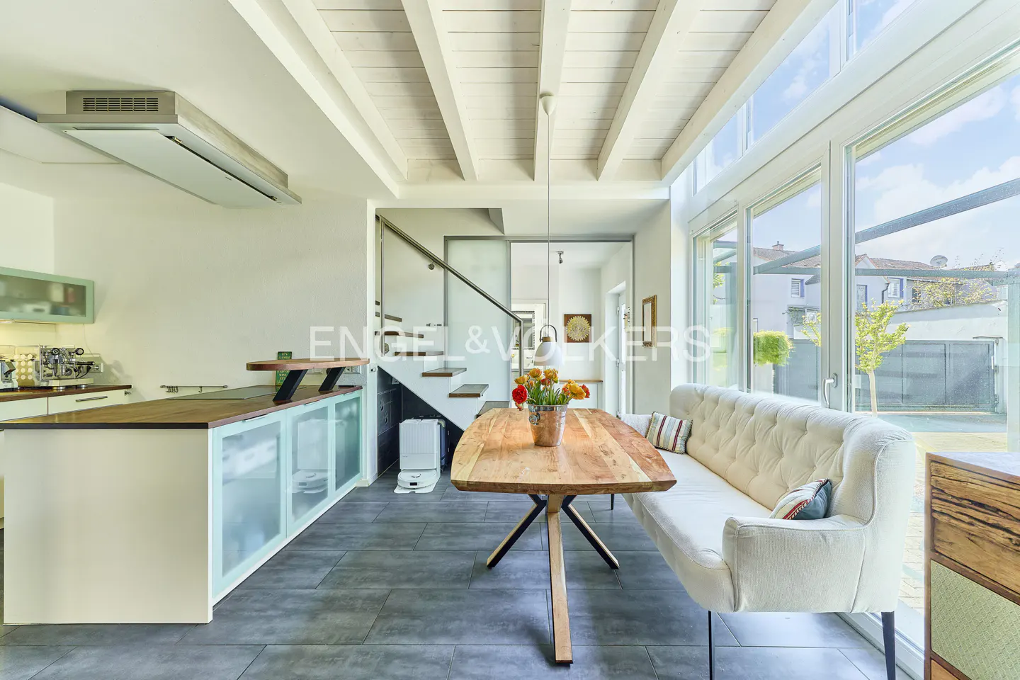 Bright, modern kitchen with white cabinets, wood countertops, and a large window. A wooden table and white sofa are in the foreground.
