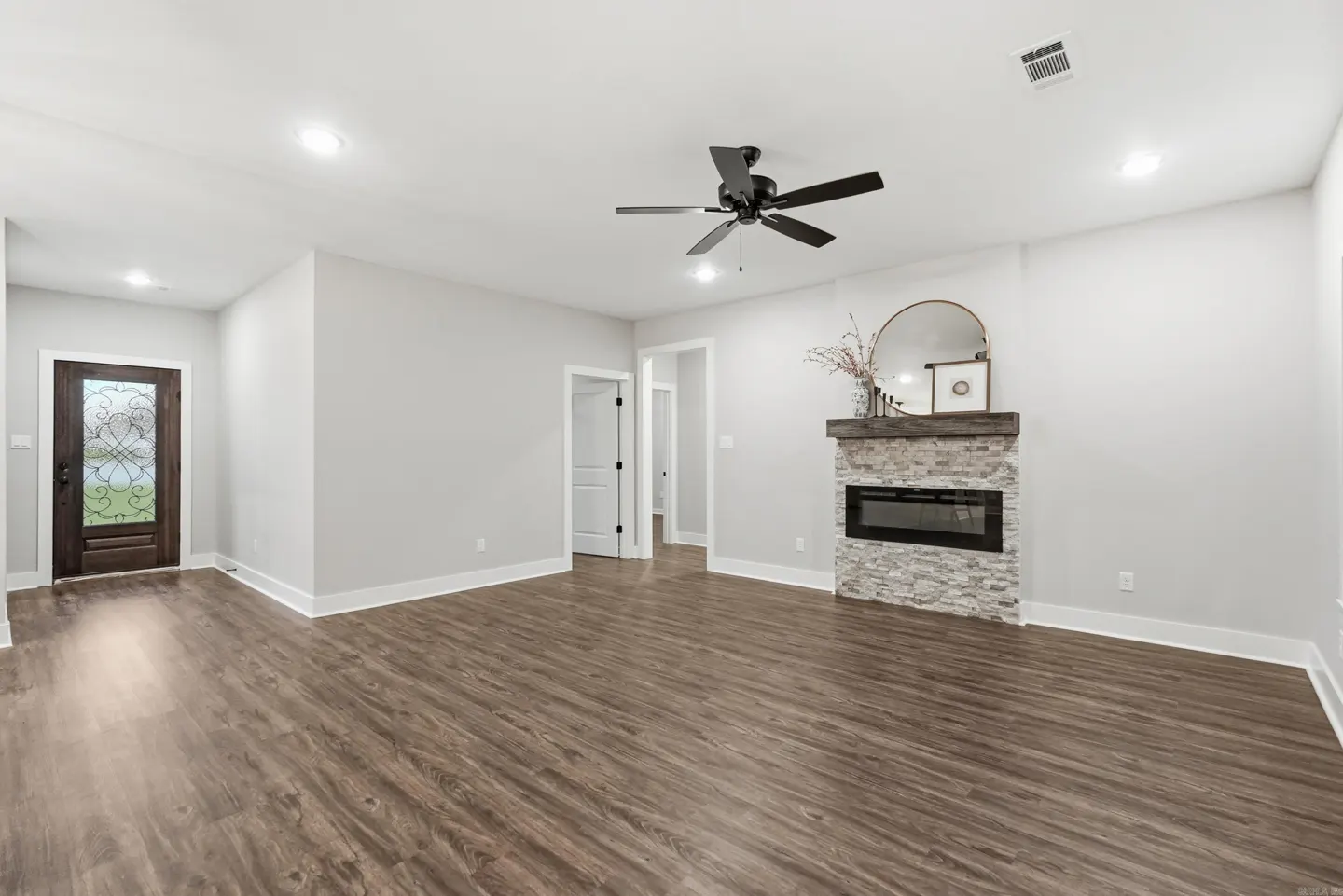 A living room with wood floors, gray walls, and a stone fireplace with a wooden mantel. A black ceiling fan hangs above.