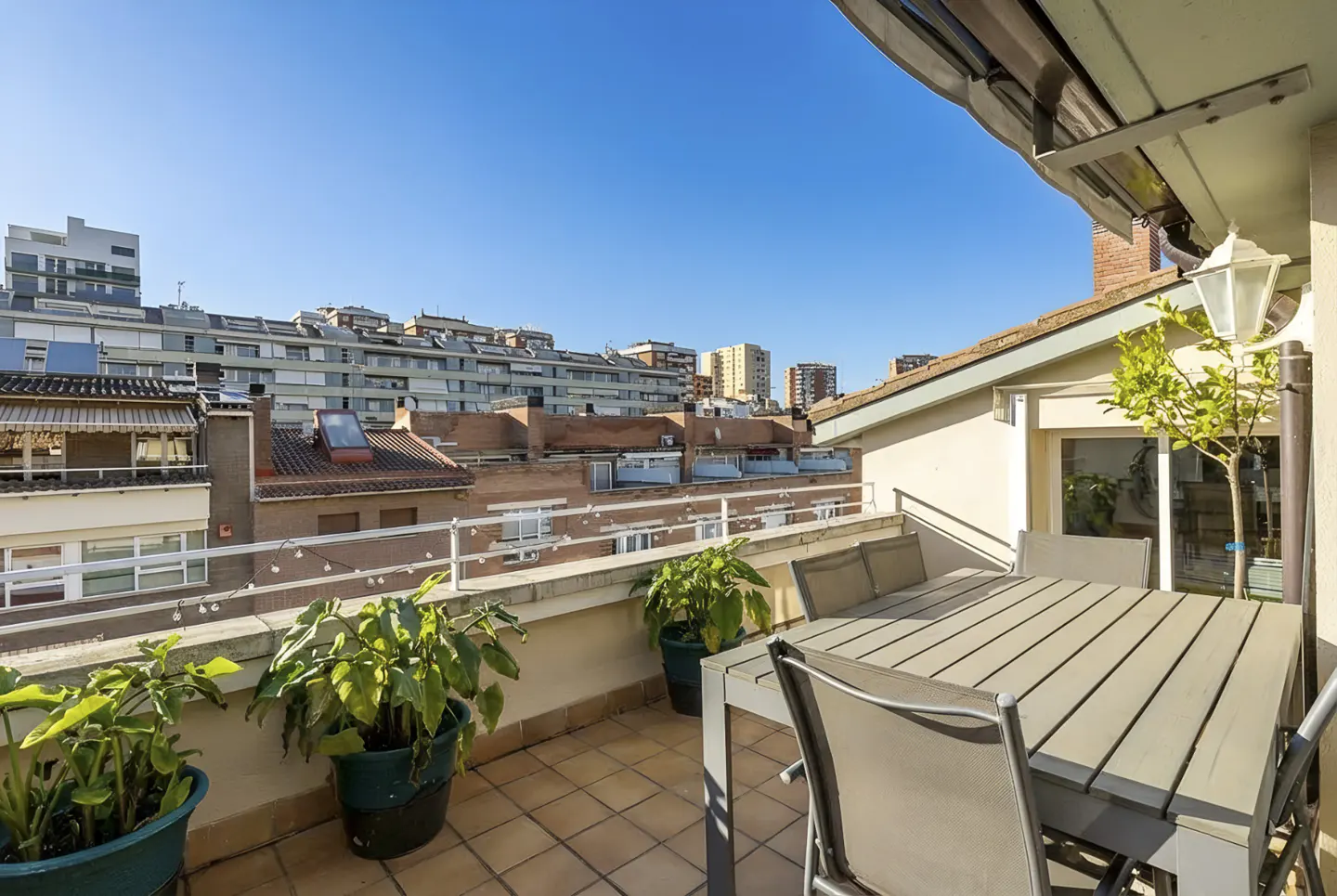 A sunny balcony with a gray table and chairs, potted plants, and a city view under a clear blue sky.