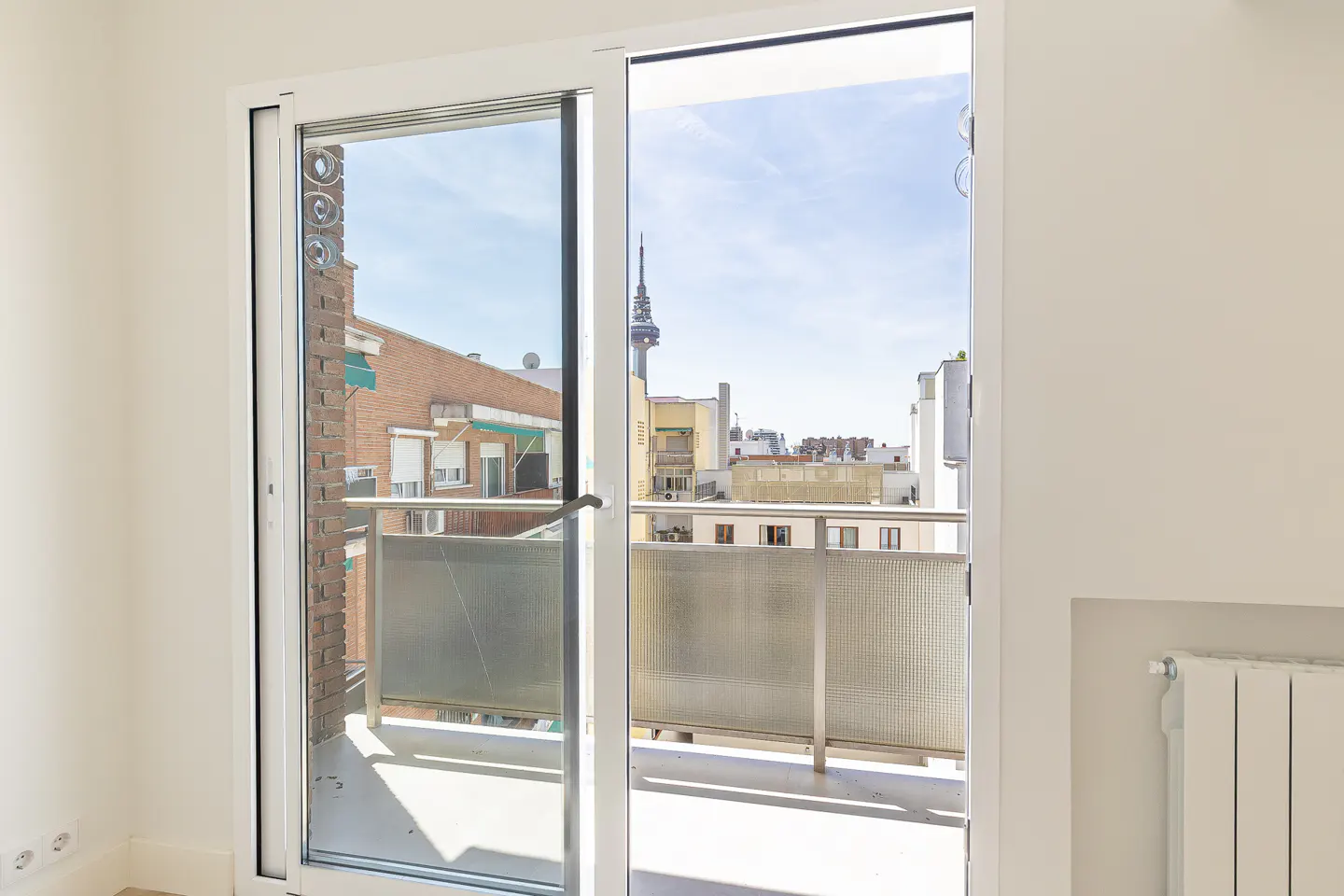 View from a white-walled room through sliding glass doors to a balcony overlooking a city with a tower.