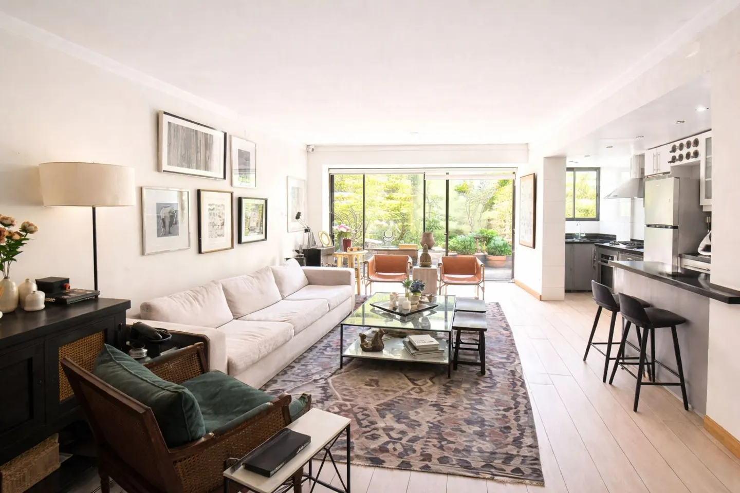 Bright living room with white sofa, patterned rug, and open kitchen. Large glass doors lead to a lush green garden.