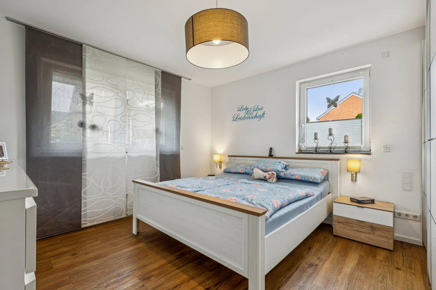 Bright bedroom with a white bed, floral bedding, and wood floors. A window and sliding curtains are on the left. A lamp hangs from the ceiling.