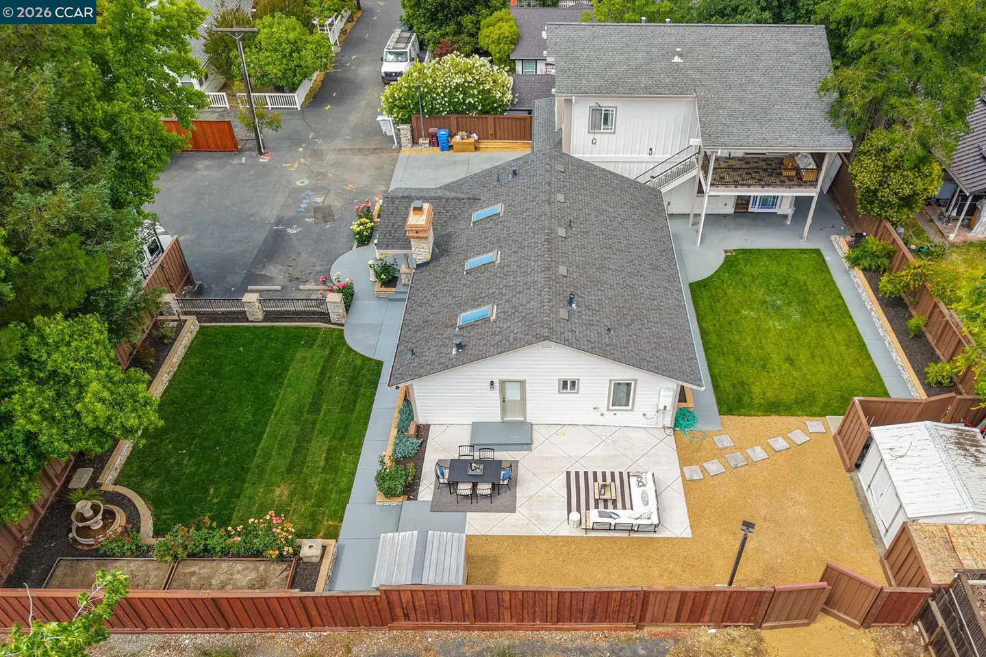 Aerial view of a white house with a gray roof, green lawns, patio furniture, and a brown wooden fence.